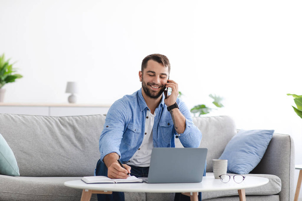 Χαμογελώντας ενήλικος Ευρωπαίος με γενειάδα κάθεται σε καναπέ εργασίας στο σπίτι εξ αποστάσεως με φορητό υπολογιστή, να μιλήσετε με τον πελάτη από το τηλέφωνο - Φωτογραφία, εικόνα