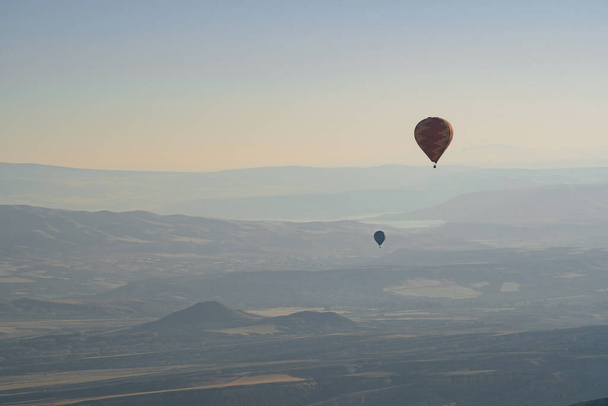 vodorovná krajina se vzdálenými horkovzdušnými balóny stoupajícími k obloze při východu slunce s krásným panoramatem údolí Cappadocia zakrytým ranní mlhou - Fotografie, Obrázek
