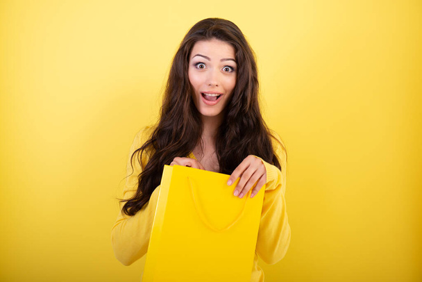 Happy surprised young woman holding shopping bag over yellow studio background. Sale, Black Friday, give-away concept - Foto, Imagen