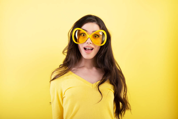 Happy smiling excited comical surprised young attractive woman with long flying hair and big funny glasses over yellow background.  - 写真・画像