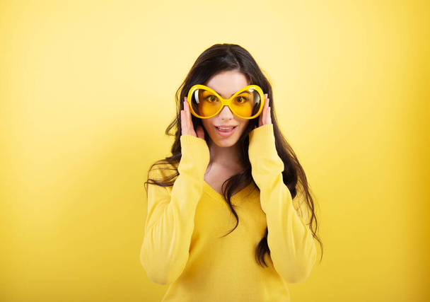 Happy smiling excited comical surprised young attractive woman with long flying hair and big funny glasses over yellow background.  - 写真・画像