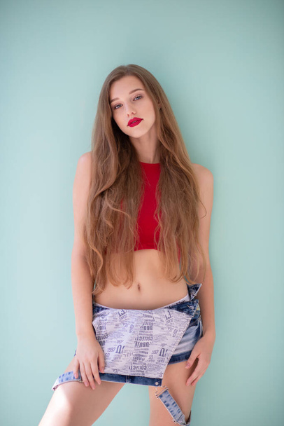 Beautiful young woman with long fair hair wearing blue jeans and red top posing indoor over light blue-green background.  - Zdjęcie, obraz