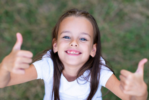 Χαριτωμένο κοριτσάκι με μακριά καστανά μαλλιά αντίχειρες πάνω από το καλοκαίρι φύση υπαίθρια. Το πορτραίτο του παιδιού με θετικές εκφράσεις. Όμορφο χαρούμενο χαμογελαστό παιδικό πρόσωπο - Φωτογραφία, εικόνα