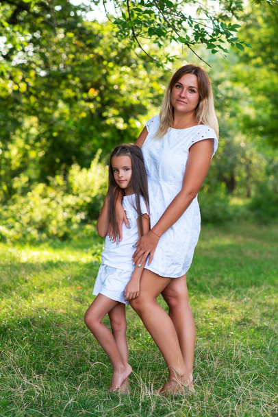 Outdoor portrait of mom and daughter wearing white clothes against summer greenery. Family walks barefooted in park. Trust, kindness, maternity, parenthood, confidence, mother's love concept.  - Foto, Imagem