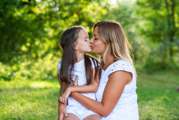 Little girl hugs kissing her mother in summer forest nature outdoor. Portrait of mom and daughter wearing white clothes against summer greenery. Family, trust, kindness, maternity, parenthood, confidence, mother's love concept.  - Фото, изображение