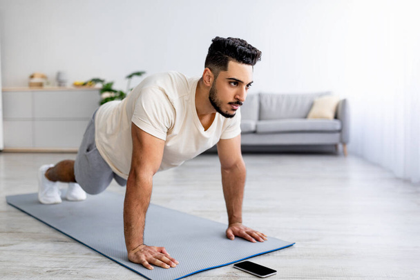 Sterkte training. Fit jonge Arabische man doen push-ups of plank thuis, na video tutorial op smartphone, vrije ruimte - Foto, afbeelding