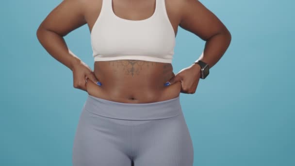 Keskiosan hidas laukaus tunnistamaton plus koko Afrikkalainen-amerikkalainen nainen ottaa ylimääräistä rasvaa puolin hänen vatsansa osoittaa sen kameran seisoo tiukka urheiluvaatteet sininen studio tausta - Materiaali, video