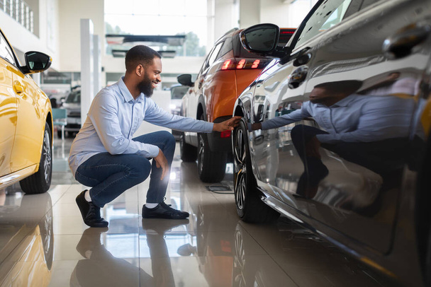 Cliente masculino negro que elige el nuevo automóvil en el centro del concesionario, examinando las llantas de la rueda - Foto, Imagen