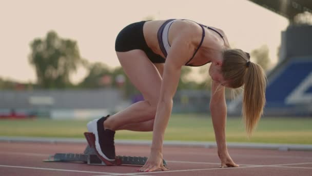 Uma jovem mulher atleta brooding aquece e se prepara para o início de uma corrida de sprint em um dia ensolarado na esteira do estádio - Filmagem, Vídeo