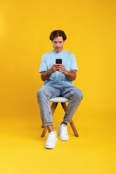 Keskittynyt kaveri käyttää kännykkää studiossa, istuu tuolilla - Valokuva, kuva