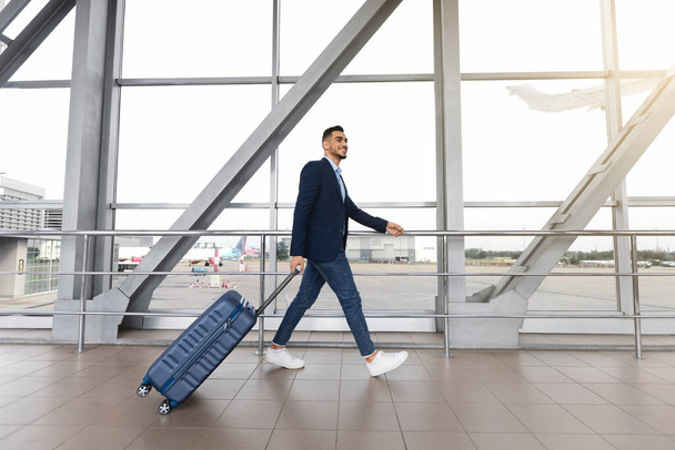 Retrato de homem de negócios árabe sorridente com mala andando perto da janela no aeroporto - Foto, Imagem