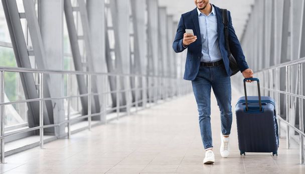 Unbekannter junger Geschäftsmann benutzt Smartphone beim Gehen mit Koffer im Flughafen - Foto, Bild