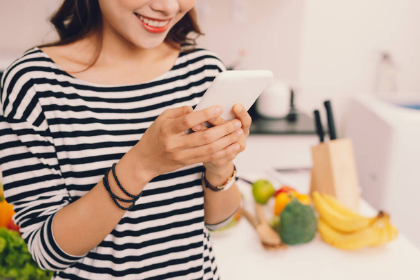 Привлекательная молодая девушка занята использованием мобильного телефона или проверкой обновлений в смартфоне во время ношения фартука на кухне  - Фото, изображение