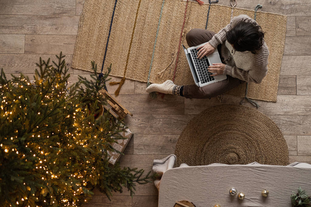 Esthétique vue du dessus de la femme assise sur le tapis et travailler sur ordinateur portable dans confortable salon confortable à la maison décoré pour Noël, fête du Nouvel An. Arbre de Noël, lumières éclatantes - Photo, image
