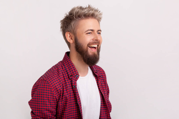 Portrait of joyful caucasian young man with beard and checkered shirt laughing, good mood and humor. Indoor studio shot isolated on gray background - Foto, immagini