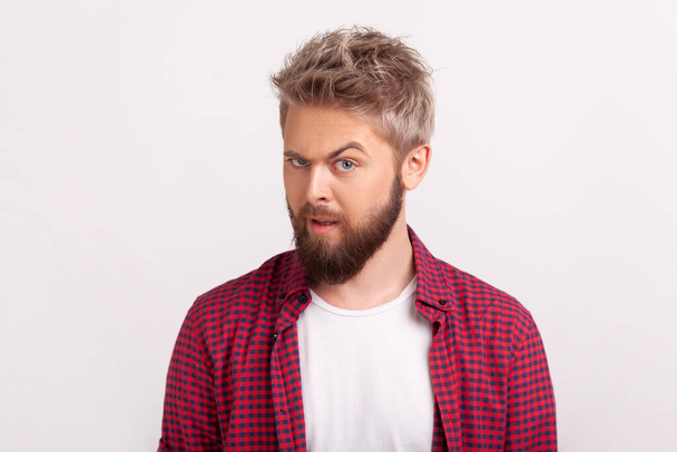Portrait of handsome doubting young man with beard wearing plaid shirt, looking at camera with raised eyebrow. Indoor studio shot isolated on gray background - Photo, Image