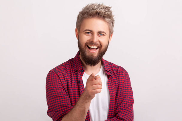 Portrait of glad bearded male in red shirt sincerely smiling at camera and gesturing hey you sign with finger. Indoor studio shot isolated on gray background - Photo, Image