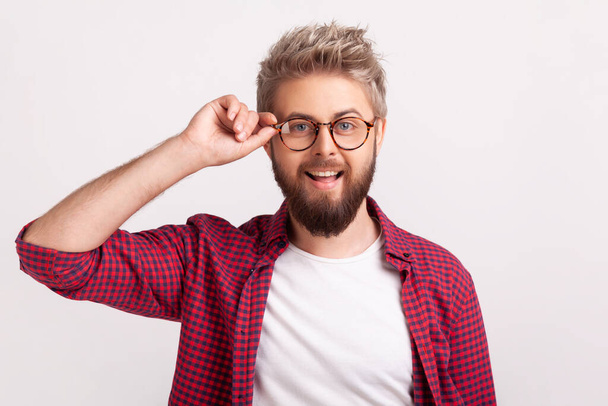 Portrait of cute male optimist in red plaid shirt touching eyeglasses and sincerely smiling at camera, positivity. Indoor studio shot isolated on gray background - Foto, Imagem