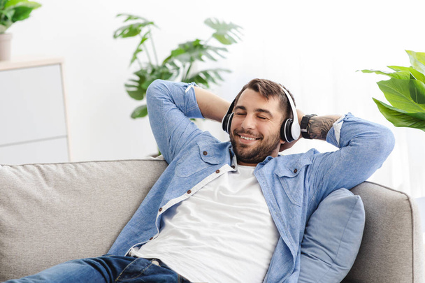 Spokojeně se usmívající dospělý Evropan s vousy a zavřenýma očima sedí na pohovce a poslouchá oblíbenou hudbu v obývacím pokoji - Fotografie, Obrázek