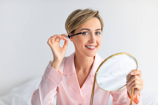 Portrait of smiling woman holding mirror applying mascara - Photo, image