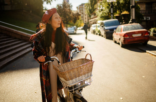 Güneşli bir günde, eski bir Avrupa şehrinin caddesinde bisikletle poz veren güzel, genç Asyalı bir kadın. - Fotoğraf, Görsel
