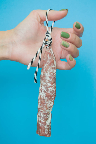 Le fuet de saucisse est accroché aux doigts des femmes. la fille garde la saucisse sur une corde. gros plan et fond bleu - Photo, image