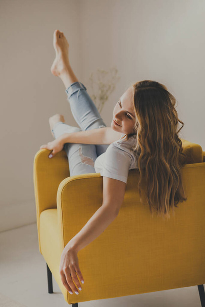 Stay at home. A young and attractive blonde is sitting in a yellow chair, with her feet on the armrest, in her apartment. Relax and happy. - Foto, immagini
