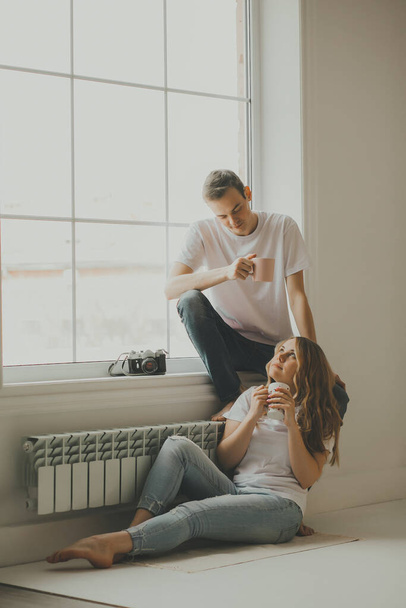 Una giovane coppia è seduta vicino alla grande finestra, riposare e bere caffè da tazze dopo un servizio fotografico, una fotocamera retrò è sul davanzale della finestra. - Foto, immagini