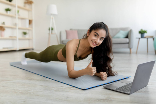 Online sports. Millennial Indian woman standing in elbow plank in front of laptop, showing thumb up gesture at home - Zdjęcie, obraz