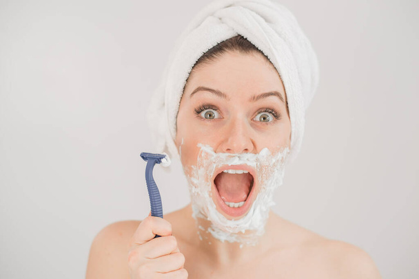 Funny portrait of a woman with shaving foam on her face holding a razor on a white background. The girl removes the mustache and beard - Photo, image