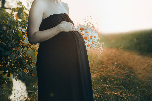 Pregnant woman hugging her tummy standing outdoors surrounded by nature. Pregnancy, expectation, motherhood concept - Valokuva, kuva