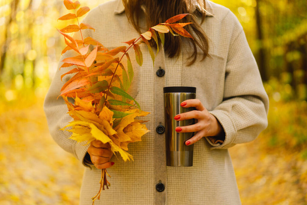 Μια γυναίκα κρατάει ένα θερμό ποτήρι και ένα μπουκέτο φθινοπωρινά φύλλα. Φθινόπωρο εξωτερική δραστηριότητα. - Φωτογραφία, εικόνα