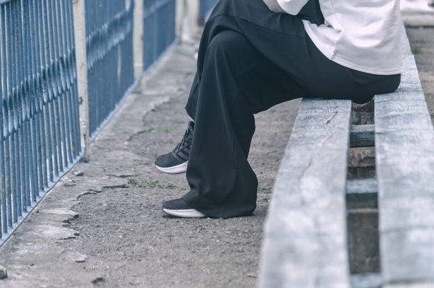 Partie inférieure de la femelle assise sur le banc. Femme en pantalon noir large, chemise blanche et chaussures de sport. Vue latérale. Concentration sélective. - Photo, image