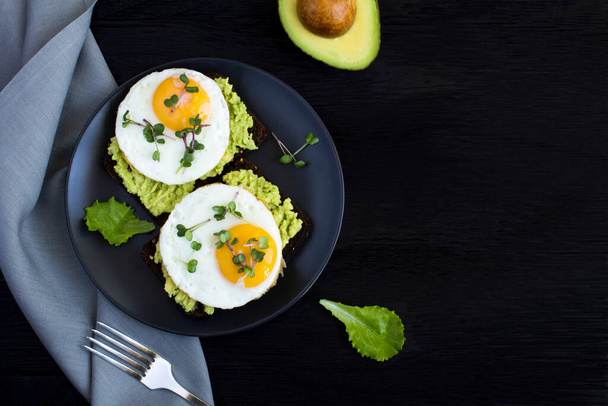Top view of avocado toasts for breakfast with rye bread, avocado puree, fried eggs and microgreen on the black  background. Copy space. - Φωτογραφία, εικόνα