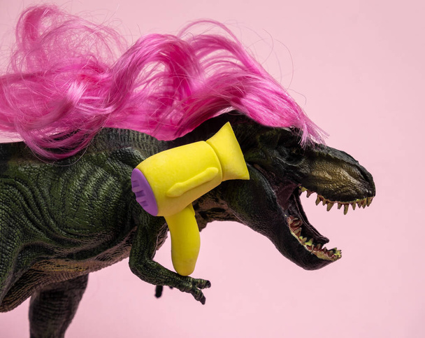 Dinosaur with long curly pink hair is holding  hairdryer. Beauty salon. Humor concept about caring for curly hair. - Photo, image