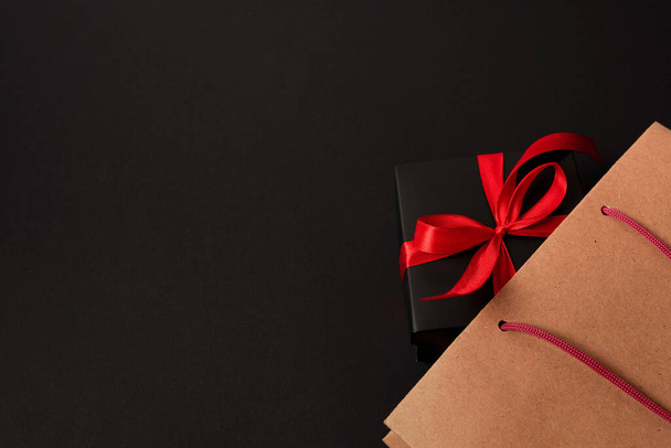 Gift box wrapped in black paper with red ribbon in paper shopping bag. Top view with copy space. - Foto, afbeelding