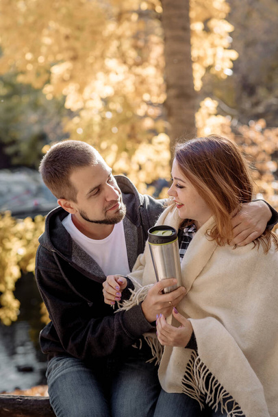 Beautiful young couple man and woman sit on a bench, drink a hot drink from a thermos and hug in a park in a leaf fall in autumn - Foto, Imagem