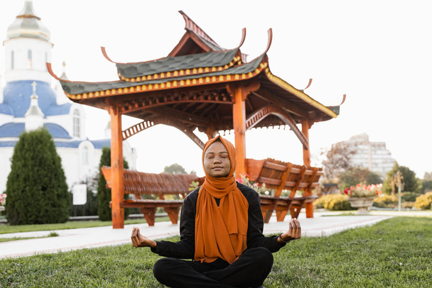 Black muslim woman meditating yoga near chinese arbor. African girl is sitting on grass and do meditation - Photo, Image