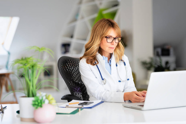 Female doctor sitting at desk and using laptop while working at doctors office - Foto, afbeelding