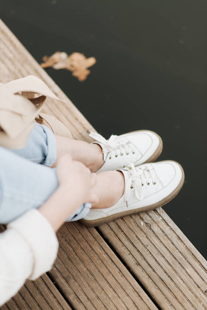 stylish female legs in white sneakers on a background of a wooden surface and water - Foto, Imagen