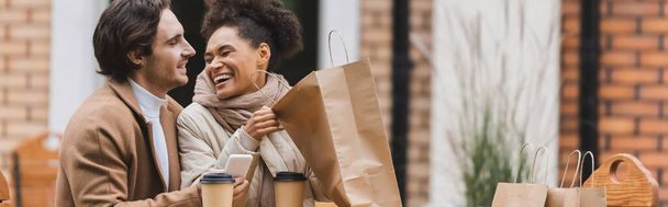 Afro-Amerikalı mutlu bir kadın alışveriş çantası tutuyor ve erkek arkadaşının yanında akıllı telefonuyla gülüyor. - Fotoğraf, Görsel