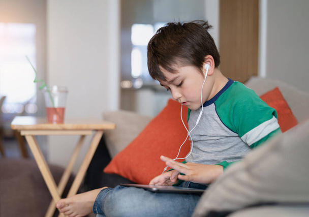 Dziecko noszące słuchawki słuchające muzyki, Młody siedzący na kanapie oglądający kreskówki lub grający w gry na tablecie, Dziecko korzystające z cyfrowej lekcji pad learning w Internecie, Nauka koncepcji edukacji online - Zdjęcie, obraz