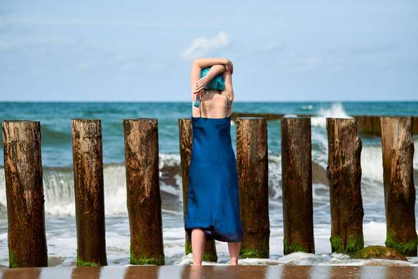 Piękna niebiesko-włosa artystka performance w ciemnoniebieskiej sukience stojąca na plaży trzymając pędzel, widok z tyłu. Sztuka współczesna abstrakcyjna, sztuka tańca w przyrodzie, sztuka nowoczesna performance outdoor - Zdjęcie, obraz