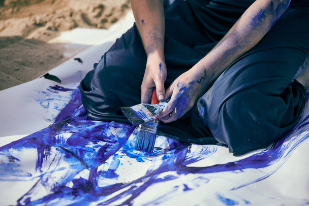 Kobieta wykonawca w ciemnoniebieskiej sukience rozmazany niebieskim malarstwem gwasz z szerokimi pociągnięciami na dużym płótnie leżącym na plaży, zbliżenie. Ekspresyjna sztuka performatywna, sztuka współczesna, outdoor - Zdjęcie, obraz