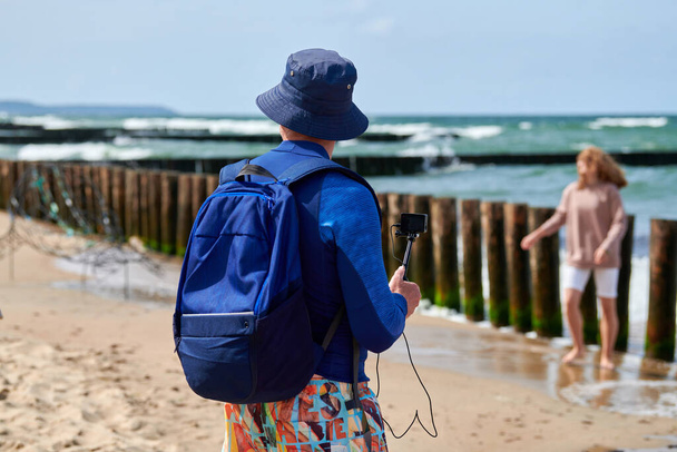 Vídeo de grabación turística masculina en cámara de video de acción, sosteniendo la cámara digital usando monópodo. Blogger en sombrero de panama, cortos multicolores y video blog de filmación de mochila en la playa en el fondo de la orilla del mar - Foto, Imagen