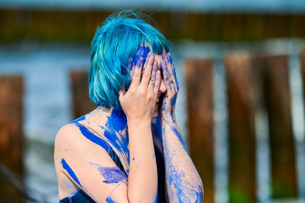 Художественная синеволосая исполнительница в темно-синем платье, размазанная гуашью индиго на своем теле на пляже, крупным планом. Креативная живопись тела, концепция боди-арта, искусство наружного перформанса - Фото, изображение