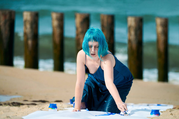 Artistic blue-haired woman performance artist in dark blue dress smeared with blue gouache painting with wide strokes on large canvas on beach. Expressive performance art, contemporary art outdoor - Photo, Image