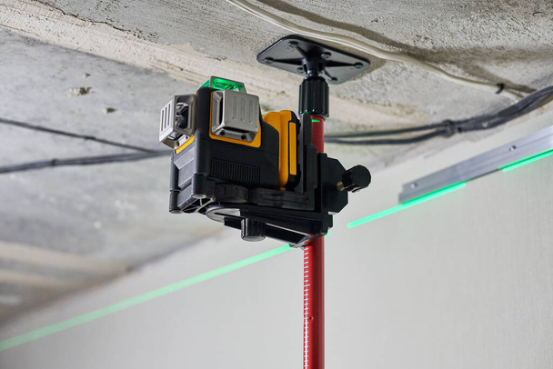 Laser level measuring tool with visible green laser beam on wall at construction site. Measuring level. Professional equipment for installing stretch ceiling. Home renovation concept, close up - Photo, Image