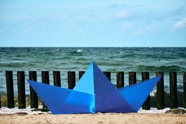 Origami blue paper boat on sandy beach for design, blue paper ship on sea shore, horizon background. Dreaming, inspiration and imagination concept. Art object at summer outdoor fine arts festival - Photo, Image