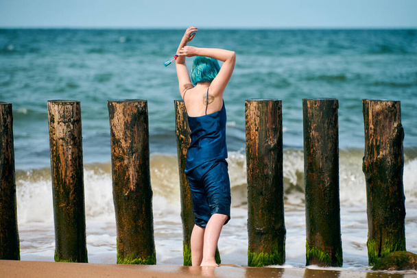 Krásná modrovlasá žena performance umělec v tmavomodrých šatech stojí na pláži drží štětec, pohled zezadu. Současné abstraktní umění, umění tance v přírodě, moderní performance outdoor - Fotografie, Obrázek